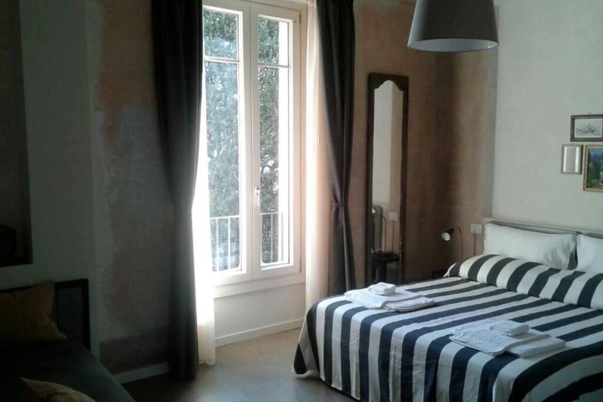 Une chambre avec une grande fenêtre, un lit avec une literie à rayures, un miroir et une lampe suspendue.