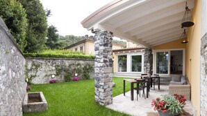 A patio area with a stone column, a dining table with chairs, and a potted plant with red flowers. - Balbianino (Tremezzina)
