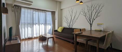 A modern living room with a wooden floor, a sofa, a dining table with chairs, an air conditioning unit, and a large window with curtains.