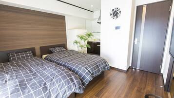 A modern hotel room with two beds, a wooden headboard, a small desk with a lamp, a potted plant, and a door.