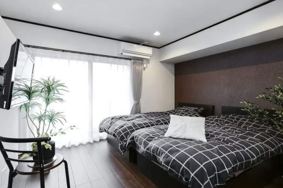 A modern hotel room with two beds, a large window, a potted plant, and a flat-screen TV.