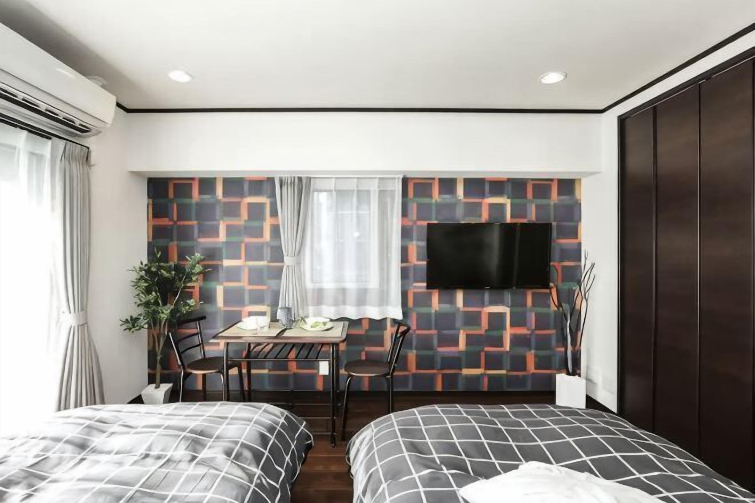 A hotel room with two beds, a desk with chairs, a TV mounted on the wall, and a window with curtains.