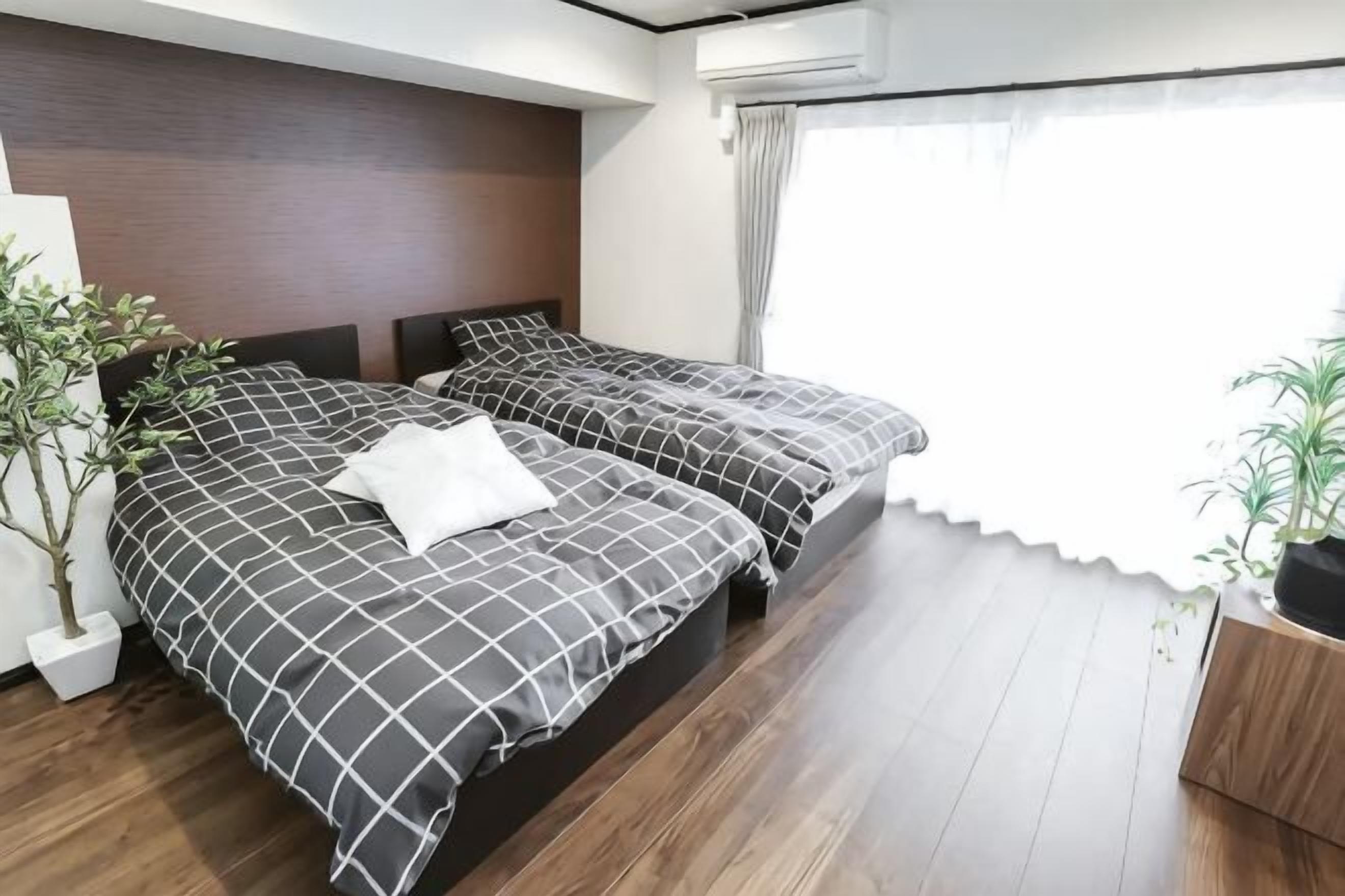 Quarto de hotel com duas camas, piso de madeira, uma janela grande com cortinas e um vaso de planta.