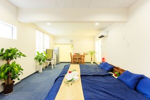 Une chambre d’hôtel avec un grand lit, un petit bureau équipé d’une télévision et d’un réfrigérateur, une table à manger avec des chaises et plusieurs plantes en pot.
