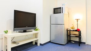 Una habitación compacta con una televisión de pantalla plana sobre un soporte blanco, un refrigerador plateado y una mesita con una lámpara y un libro.