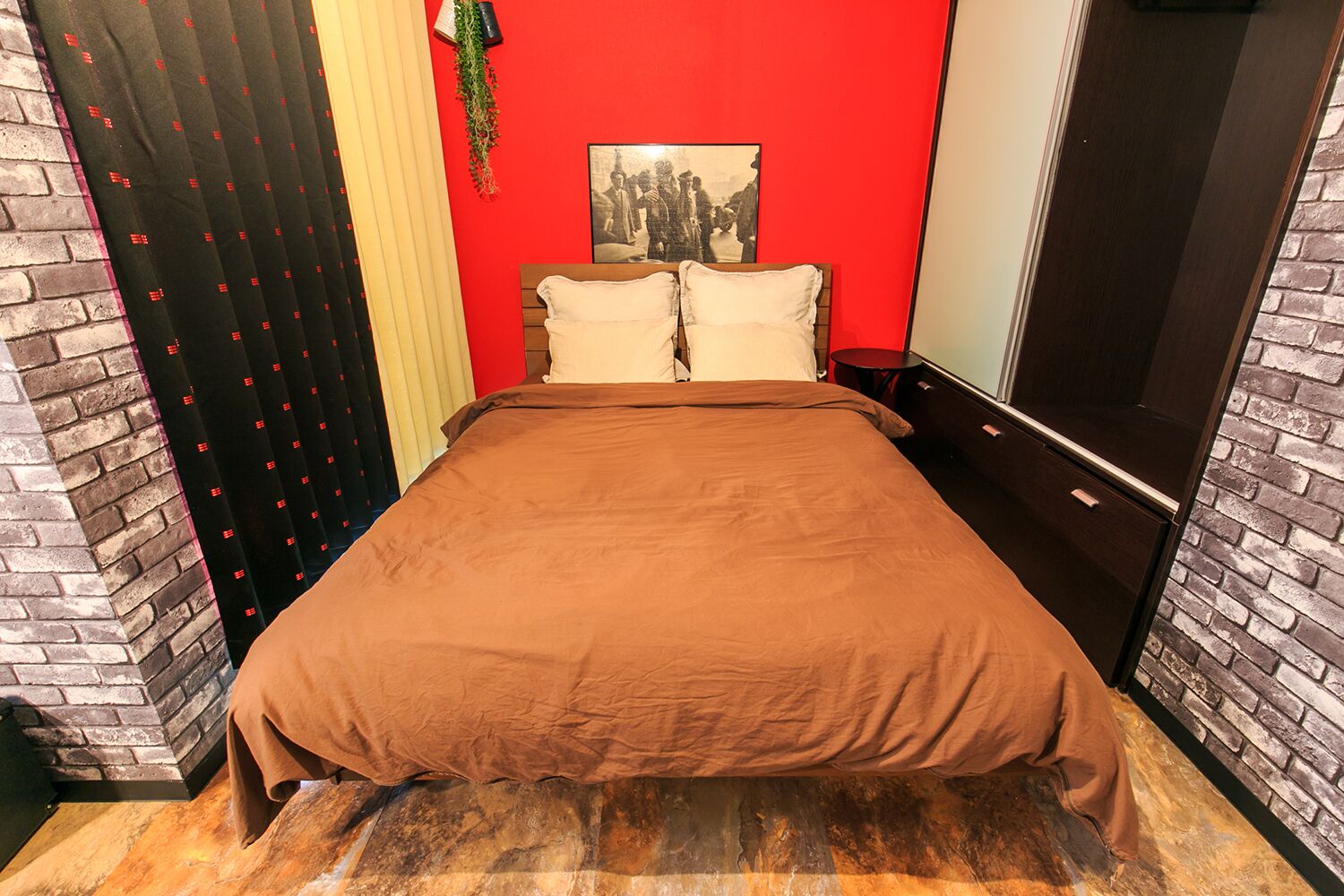 Ein Schlafzimmer mit einer Ziegelwand, einer roten Akzentwand, einem Bett mit brauner Bettdecke und einem schwarzen und weißen Bilderrahmen an der Wand.