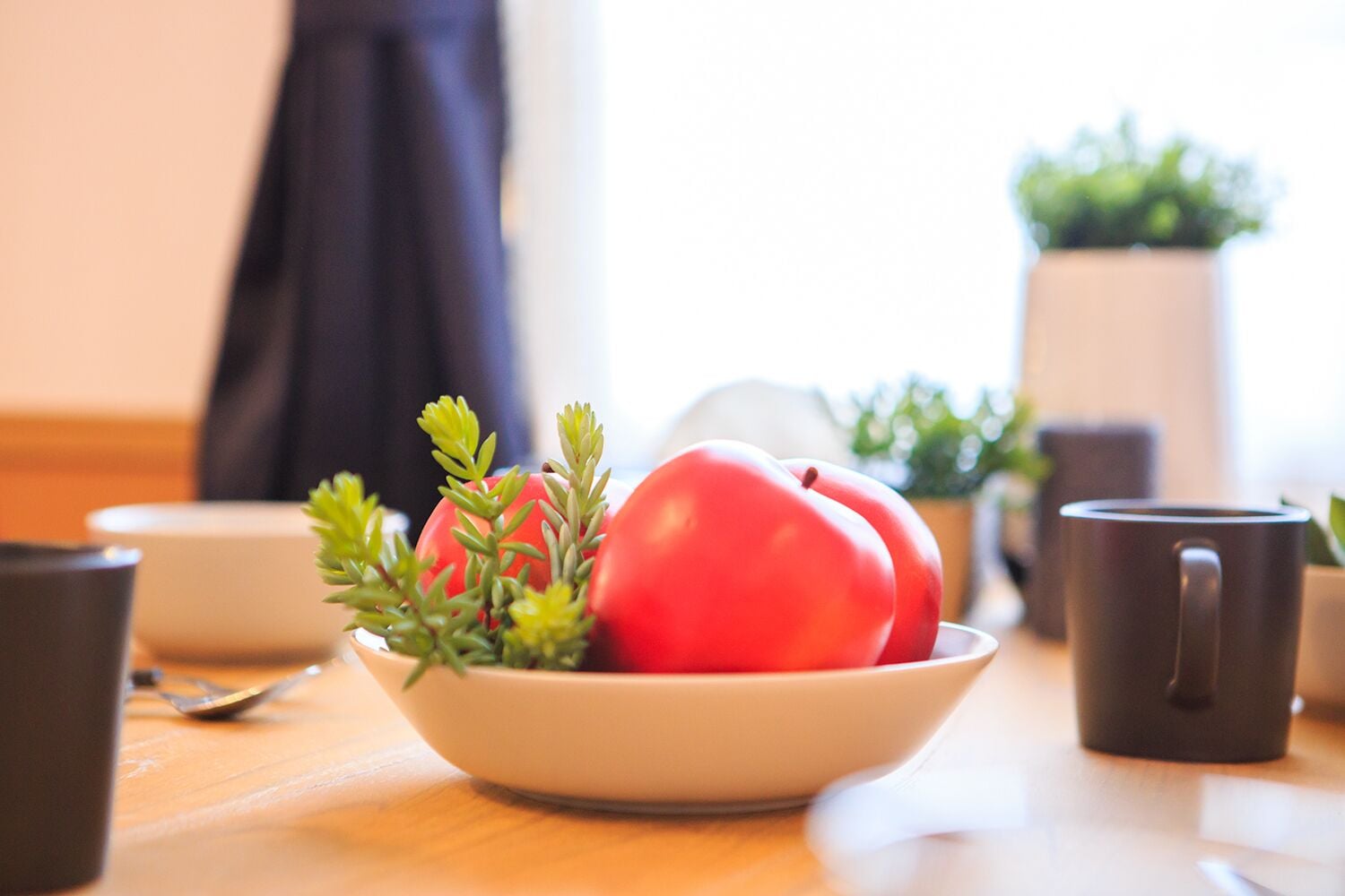 A bowl of red apples and green herbs on a wooden table with a dark mug and a white bowl in the background.