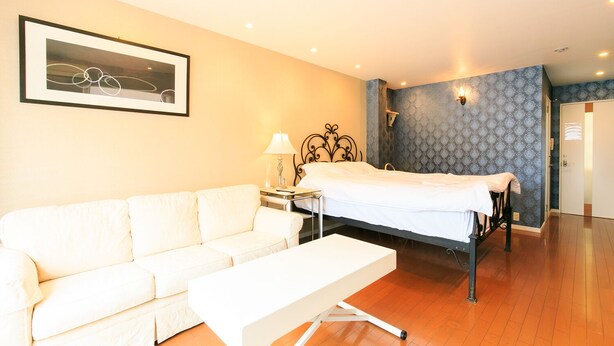A hotel room with a white sofa, a bed with a white bedspread, a bedside table with a lamp, and a framed picture on the wall. - Casa de Marzo (Nagoya)