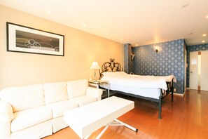 Habitación de hotel con sofá blanco, cama con cubrecama blanco, mesita de noche con lámpara y un cuadro enmarcado en la pared.