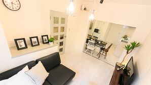 Un salon moderne avec un canapé noir, un coin repas avec une chaise blanche, un meuble pour la télévision et un grand miroir qui reflète le coin repas.