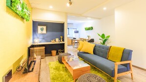 A modern living room with a grey sofa, yellow cushions, a wooden coffee table, and a green rug. There is a kitchen in the background with wooden cabinets and a dining area. - ORIGAMI STAY -Taishocho- (Nagoya)