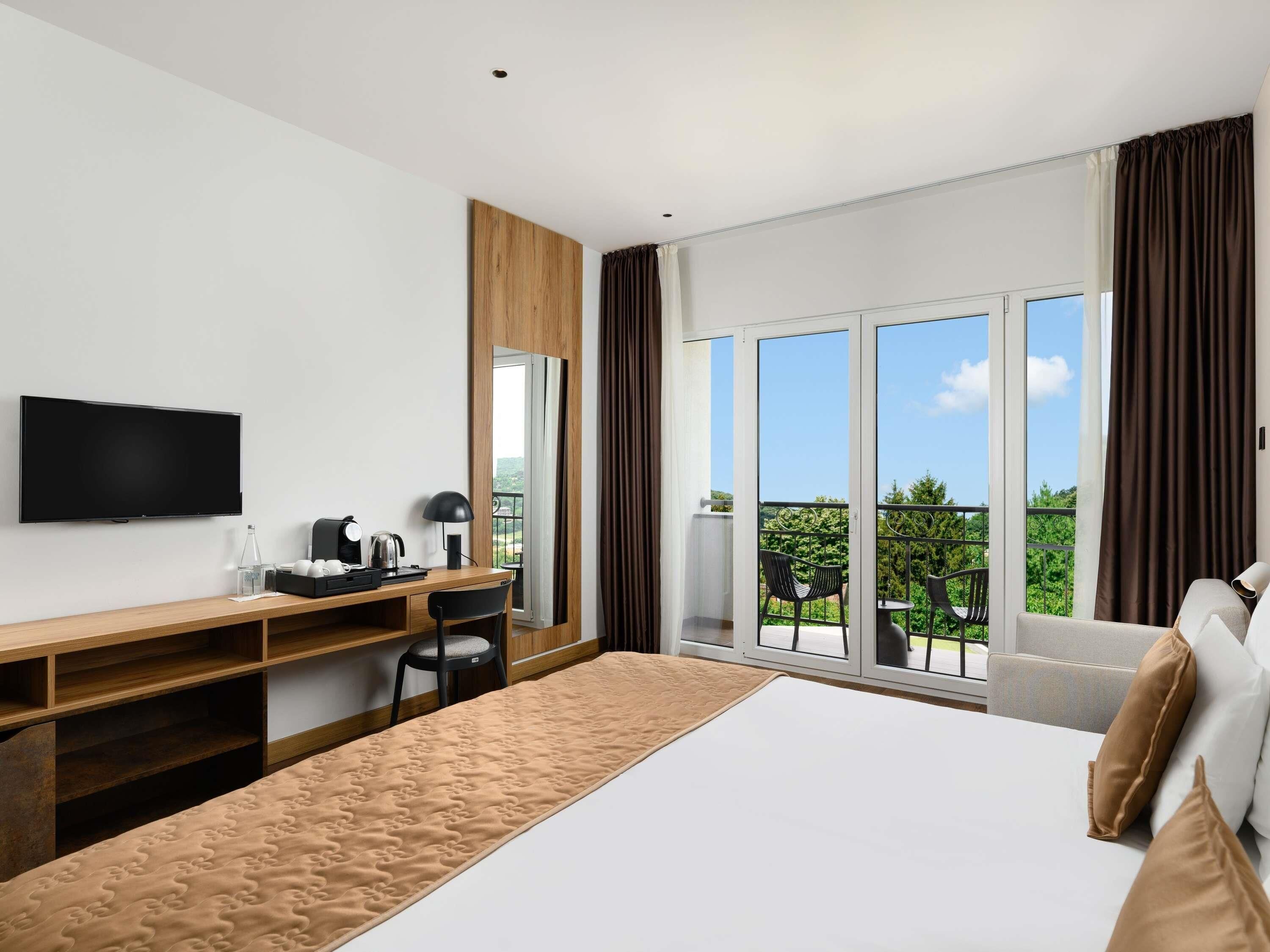A hotel room with a bed, a desk, a TV, and a balcony with outdoor furniture.