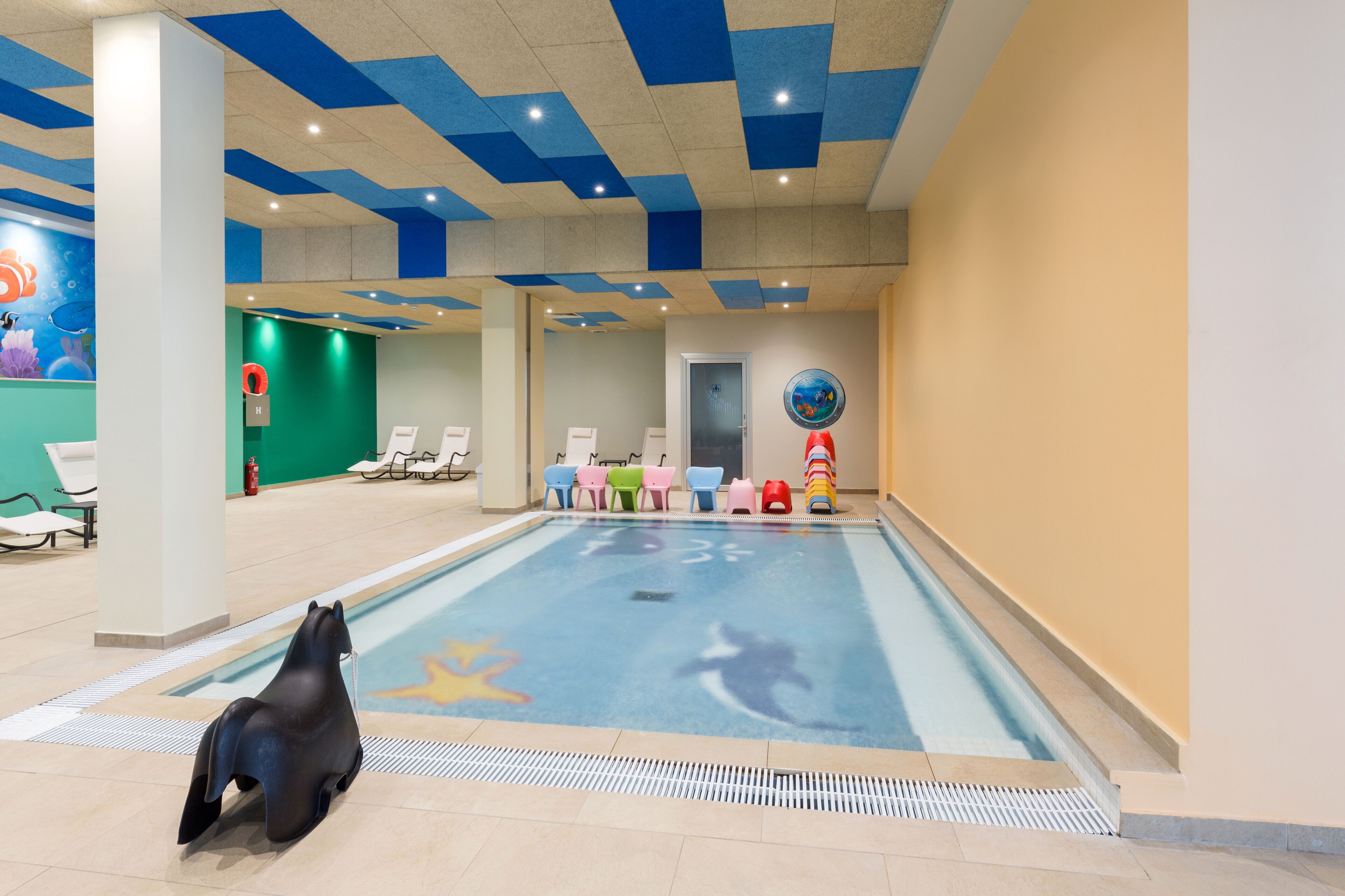 Una piscina interior con un mural de delfines, sillas de colores y una escultura de una foca.