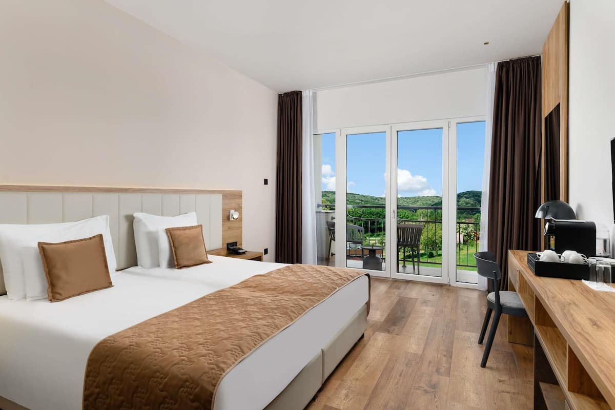 A hotel room with a bed, a desk, a chair, a nightstand, and a view of the outdoors.