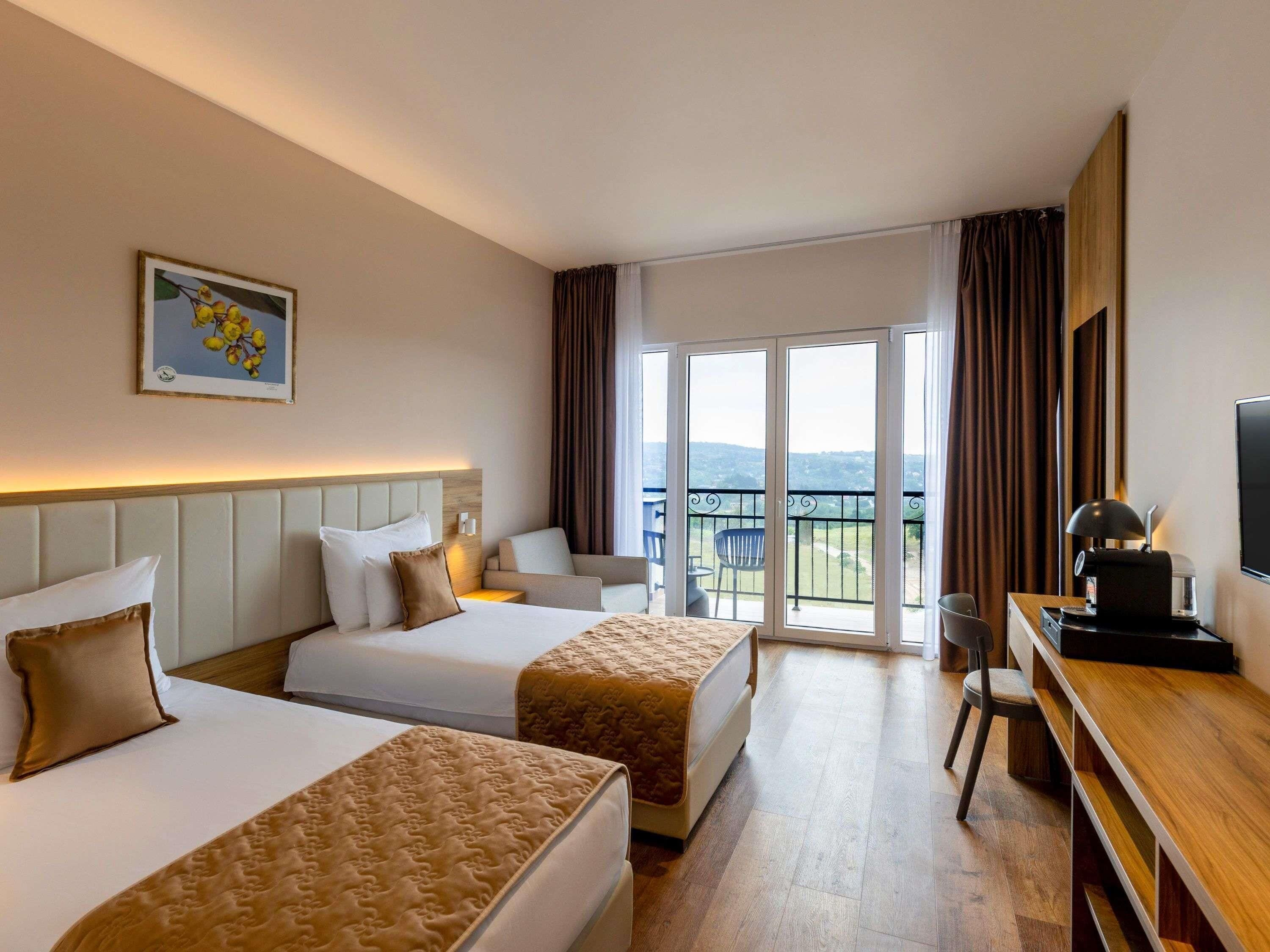 A hotel room with two beds, a desk with a lamp, a TV, and a balcony with a view.
