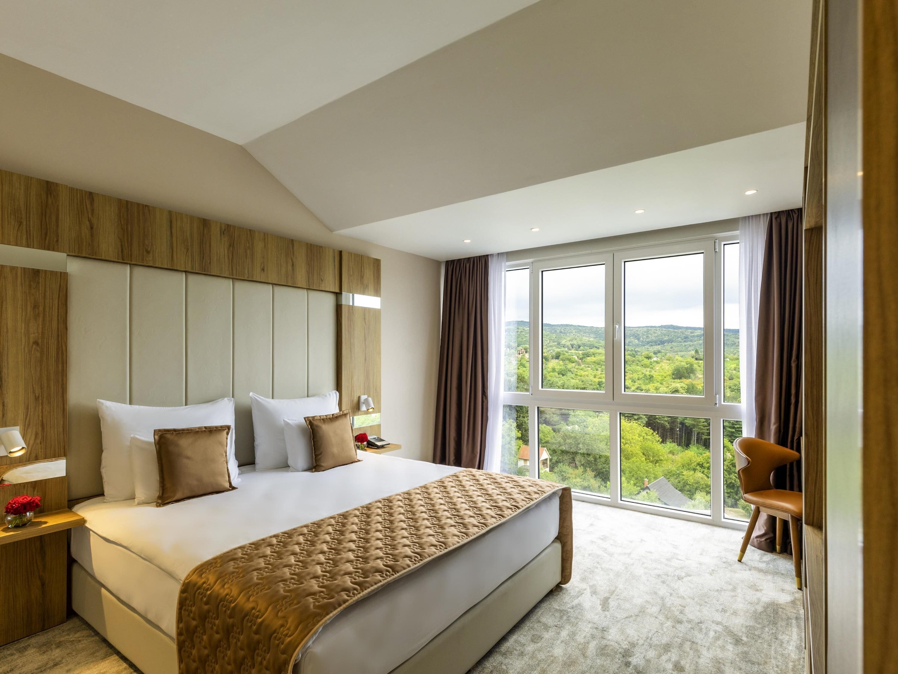 A modern hotel room with a large bed, a wooden headboard, a view of greenery, and a seating area with a chair.