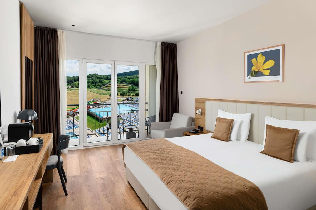 A hotel room with a bed, a desk, a chair, a nightstand, a lamp, and a view of a pool and greenery.