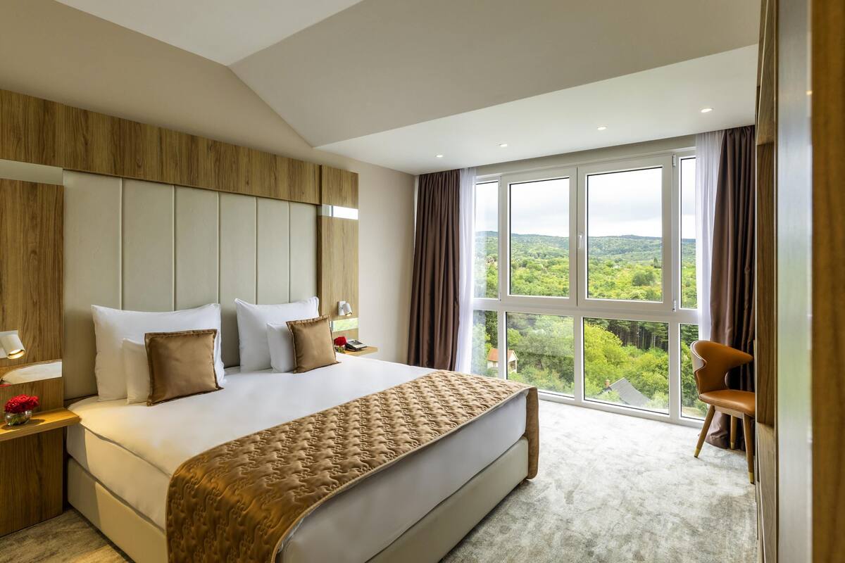 A modern hotel room with a large bed, a wooden headboard, a view of greenery, and a seating area with a chair.