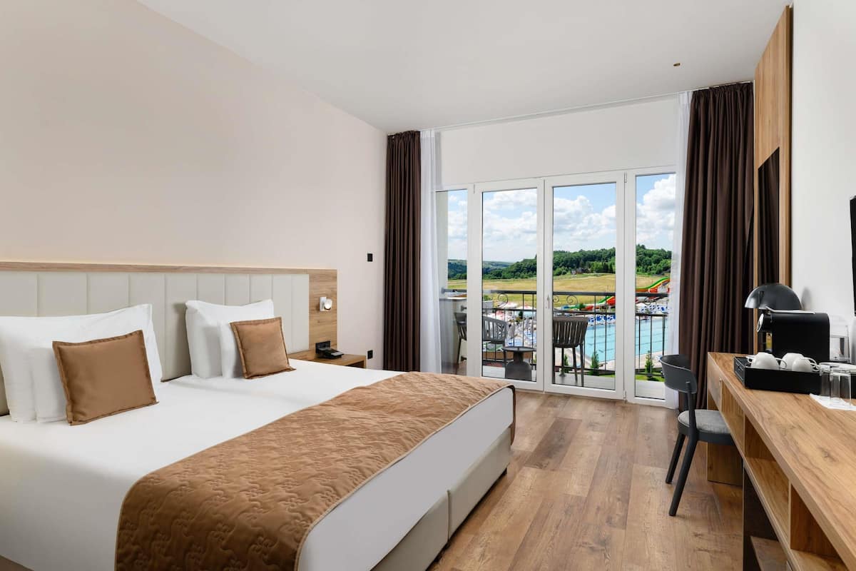 A hotel room with a bed, a desk, a chair, a TV, and a view of a swimming pool and greenery.