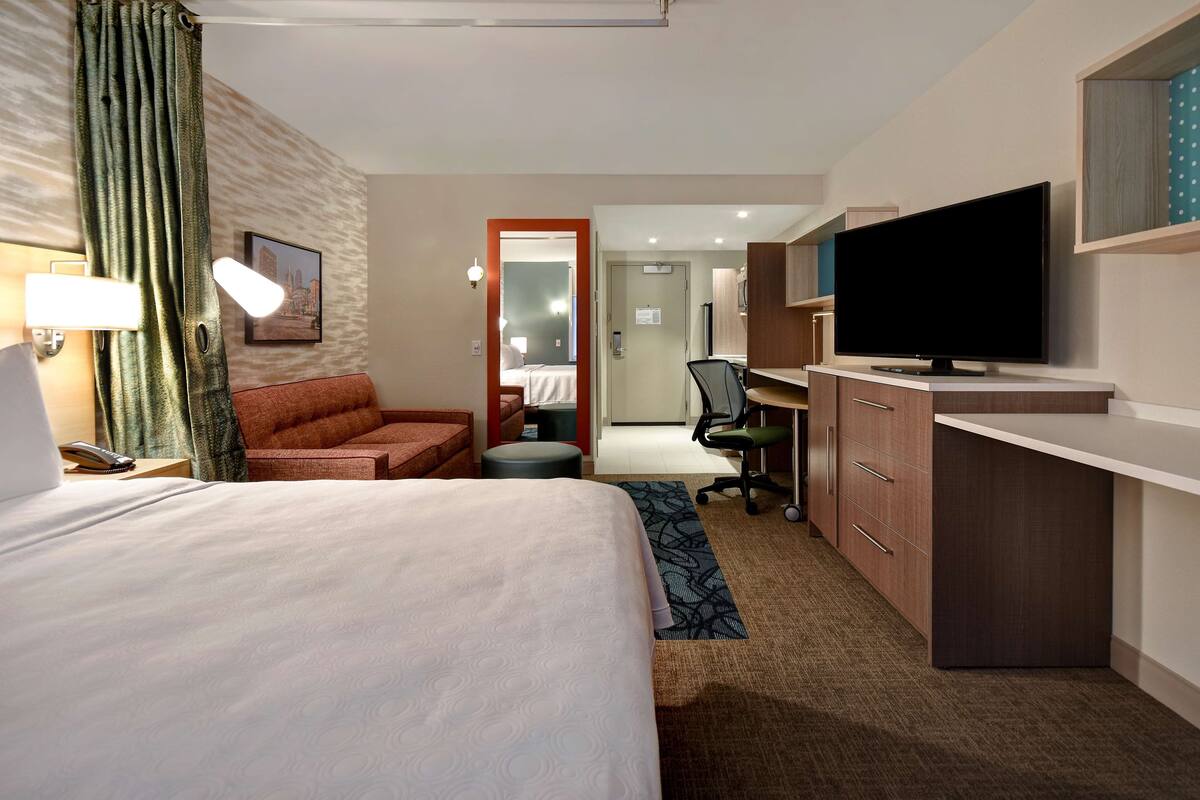 A hotel room with a bed, a desk with a TV, a chair, a desk lamp, and a window with curtains.