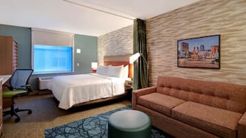 Una habitación de hotel con cama, sofá, escritorio con silla, lámpara de pie, ventana con persianas y un cuadro de paisaje urbano en la pared.