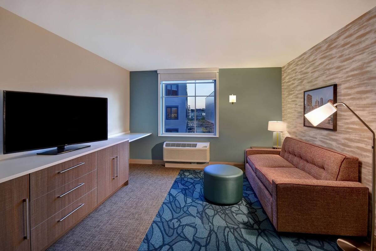 A hotel room with a flat-screen TV on a wooden cabinet, a sofa, a patterned rug, a window with a city view, and a wall-mounted lamp.