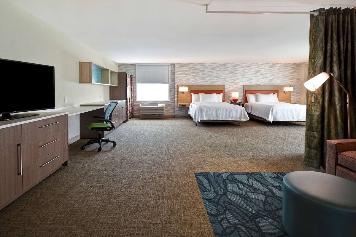 A hotel room with a TV, desk, office chair, two beds, and a small seating area.