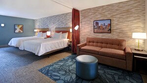 Habitación de hotel con dos camas, un sofá, un reposapiés redondo, una pintura de un paisaje urbano y una alfombra estampada.