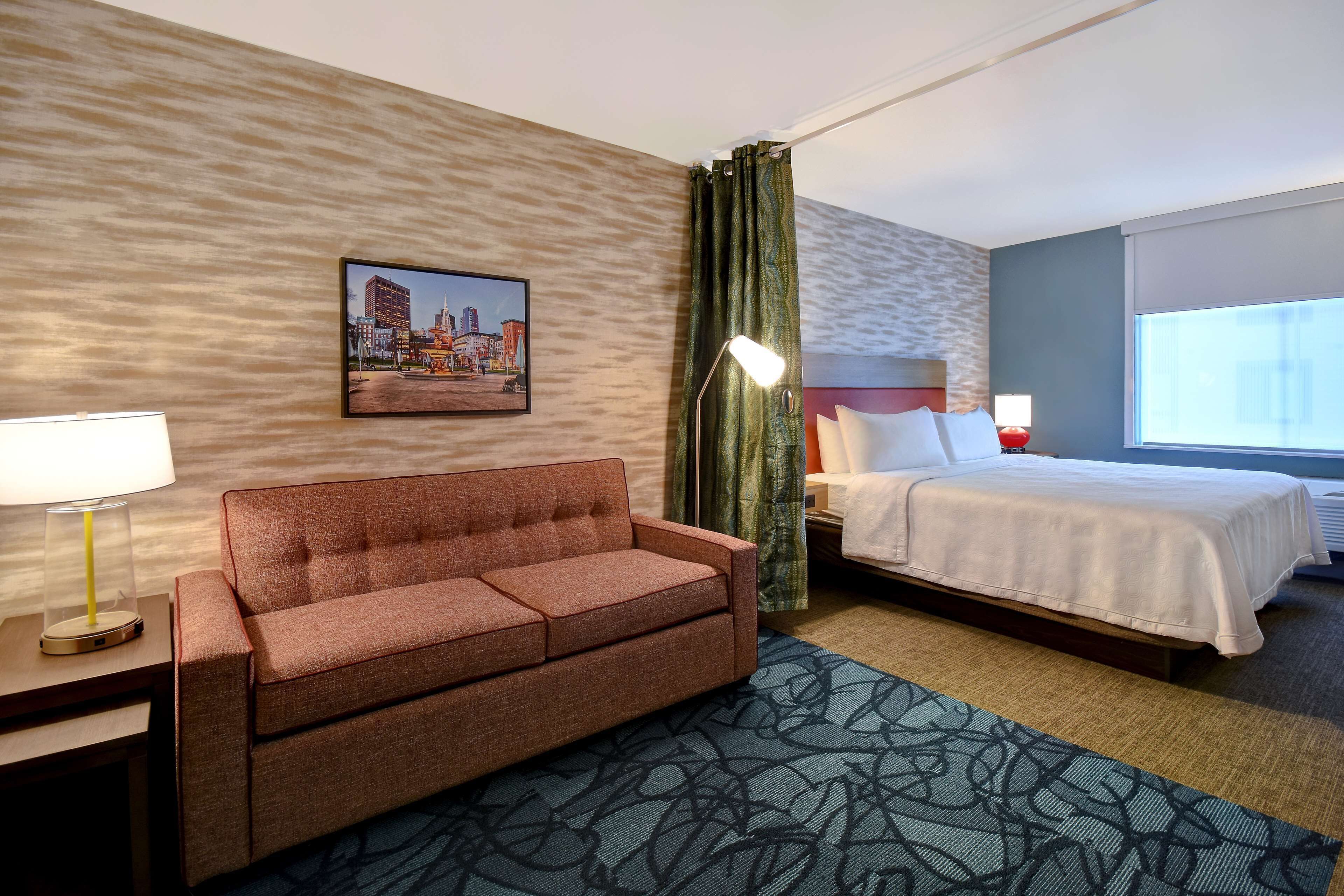 A hotel room with a sofa, a bed, a lamp, a picture on the wall, and a window.