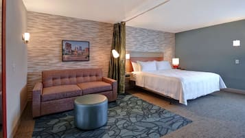 A hotel room with a bed, a sofa, a small round ottoman, a patterned rug, and a cityscape painting on the wall.