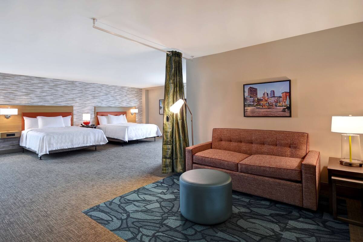 A hotel room with two beds, a sofa, a round ottoman, and a cityscape painting on the wall.