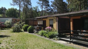 A small, single-story building with a covered patio area, surrounded by a well-maintained lawn and various shrubs.