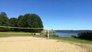 A sandy beach area with a volleyball net, a wooden bench, and a water body in the background.