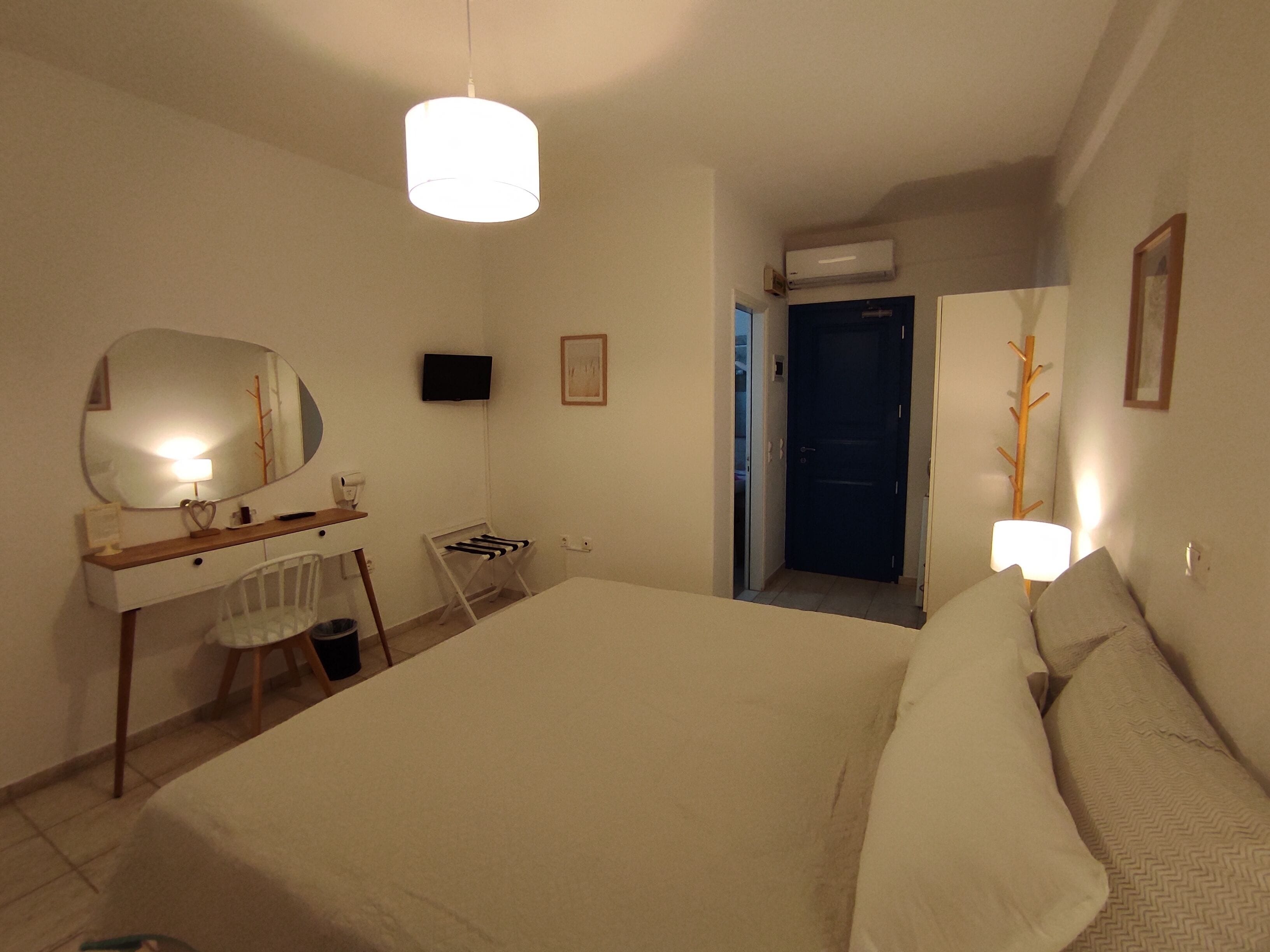 Une chambre à coucher comprenant un lit, un bureau, une chaise, un miroir, une télévision et une porte donnant sur une autre pièce.