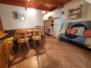 A cozy living room with a sofa, wooden chairs, a dining table, and a kitchen area with a stove and refrigerator. - GARDEN OF OLEANDERS - a stone's throw from the Sea (Menton)