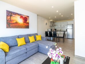 A modern living room with a grey sofa, yellow cushions, and a painting of a sunset. A dining area with a table and chairs is visible in the background.