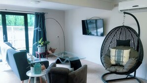 Ein Wohnzimmer mit einer Couch, Sesseln, einem Hängesessel, einem Tisch mit einer Topfpflanze und einem Fernseher an der Wand.