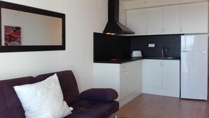 Um espaço de estar compacto com sofá roxo, ventilador de teto preto, geladeira branca e cozinha com armários brancos e bancada preta.