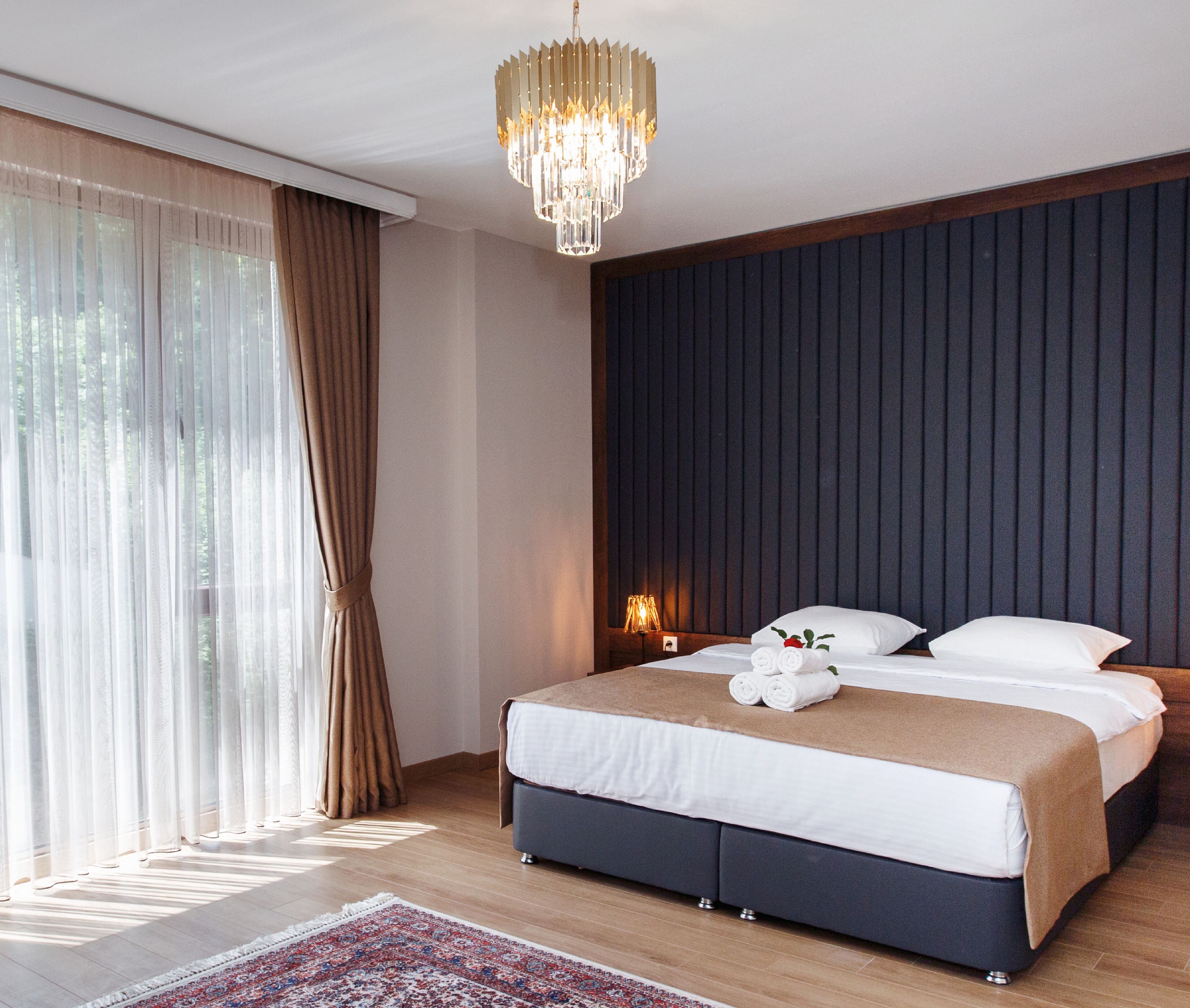 Habitación de hotel moderna con una cama grande, un candelabro y vistas a zonas verdes a través de cortinas translúcidas.