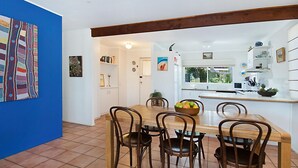 A dining area with a wooden table and chairs, a kitchen in the background, and a blue wall with a colorful artwork.