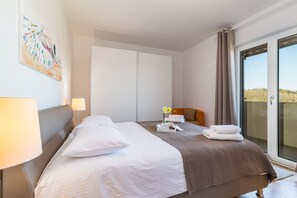 A hotel room with a large bed, a bedside table with a lamp, a small table with towels and flowers, a sliding door to a balcony, and a painting on the wall.