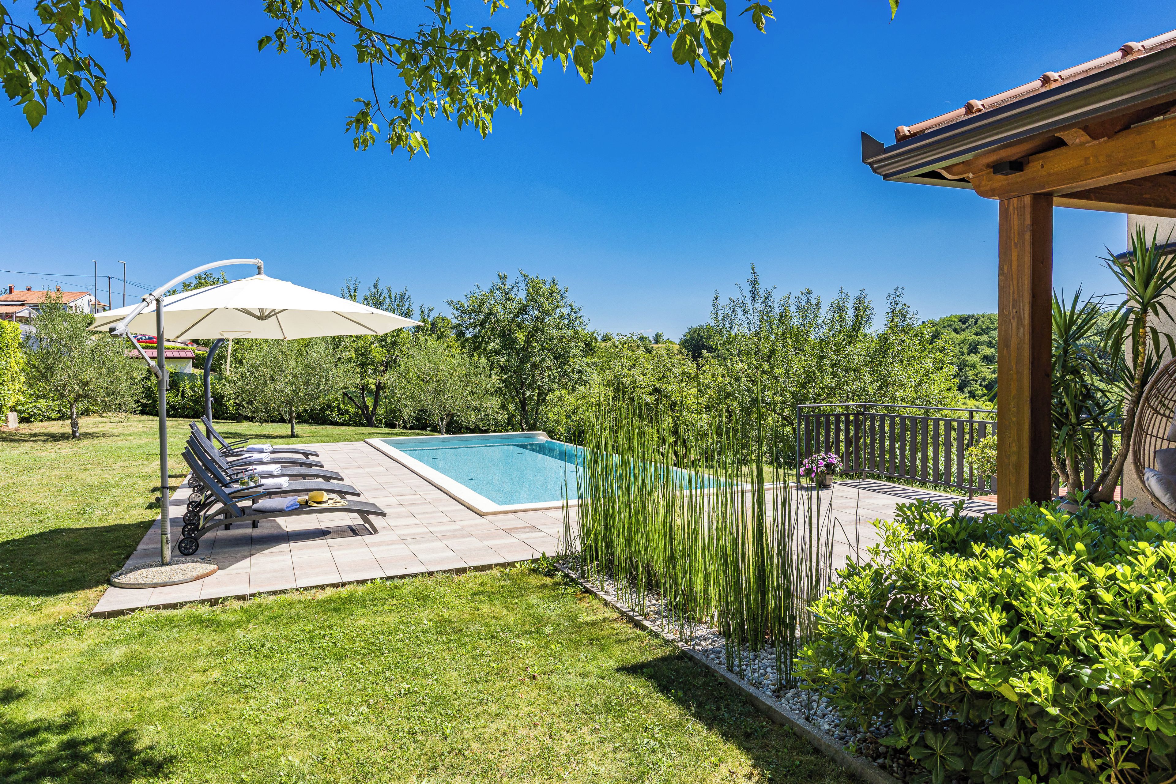 Une piscine entourée de verdure, avec des chaises longues et un parasol, sous un ciel dégagé.