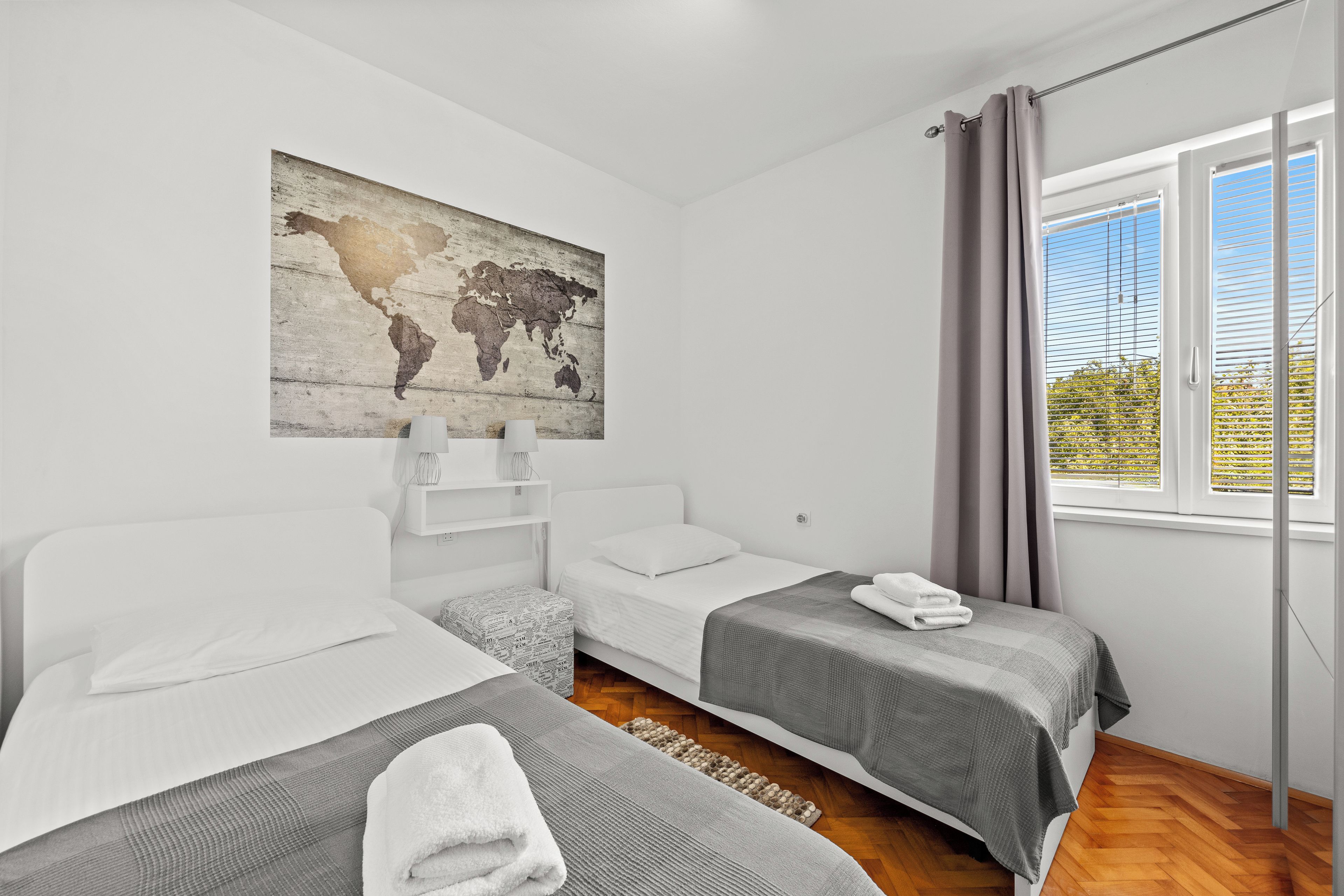 A hotel room with two beds, a world map artwork, a window with curtains, and a view of greenery outside.