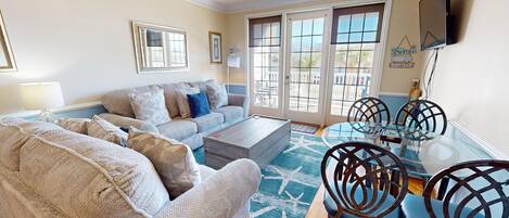 A living room with a sofa, coffee table, and chairs, a dining table with chairs, and a TV mounted on the wall.