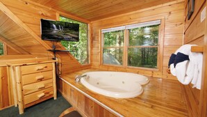 Un baño de cabaña de madera con una bañera grande, armarios de madera, un televisor montado en la pared y una ventana con vistas a los árboles.