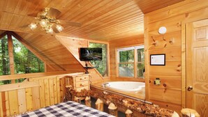 Una cabaña de madera con una cama a cuadros, un ventilador de techo, un televisor, una bañera y un ventanal grande.