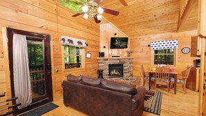 Interior acogedor de una cabaña con chimenea de piedra, sofá de cuero, zona de comedor y televisor sobre la chimenea.