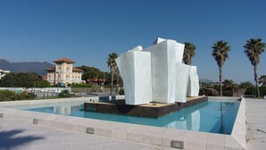 Een modern zwembadgedeelte met een opvallend witte sculptuur in het midden, omringd door palmbomen en een helderblauwe lucht.