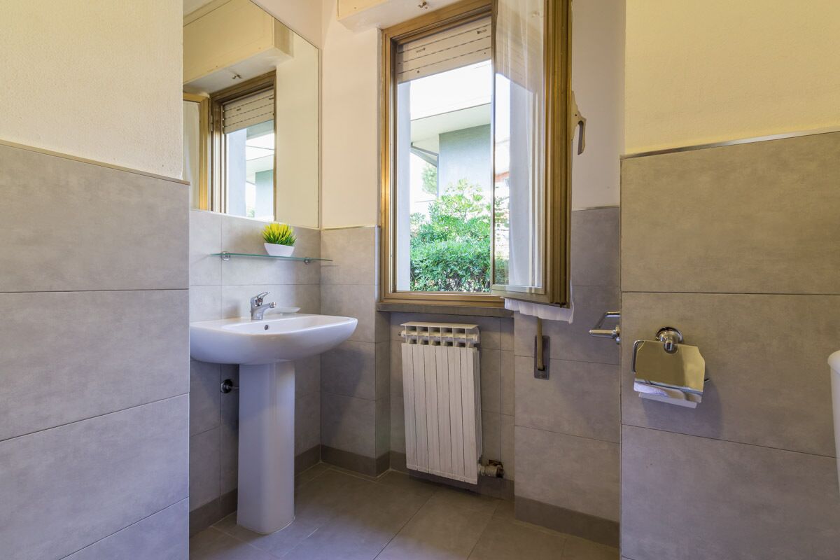 A bathroom with a pedestal sink, a window with a view of greenery, a radiator, and a towel rack.