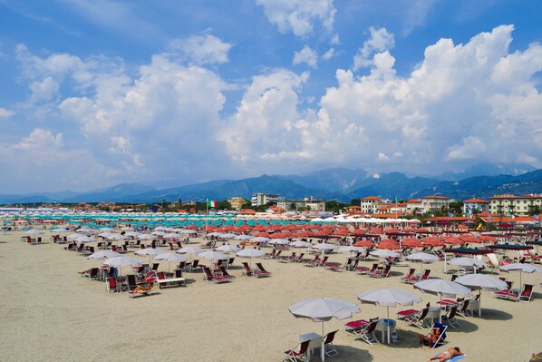 A beach with rows of sunbeds and umbrellas, a clear sky, and mountains in the background.
